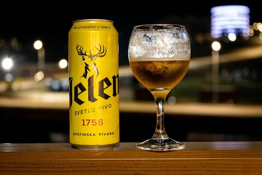 Closeup shot of a can of Jelen beer with a glass of beer on a wooden balcony railing at night