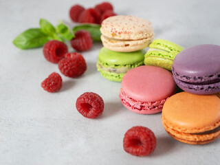 a lot of multi-colored macarons, macaroons and raspberries on a gray background with copy space. French dessert and red berries.