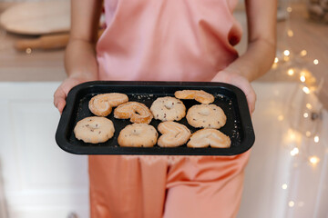 a girl with dark straight hair in light pink pajamas sits on a chair in a kitchen interior decorated in a New Year's style with a Christmas tree and various decorations and holds a baking sheet 