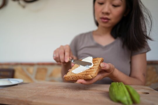 East Asian Woman Spreading Cream Cheese On Toasted Bread To Make Avocado Toast.