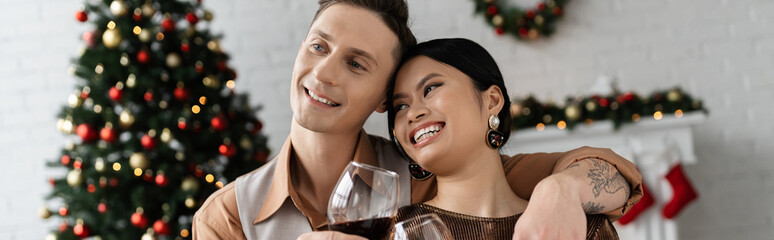 happy interracial couple clinking wine glasses near Christmas tree on blurred background, banner