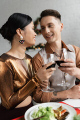 joyful multiethnic couple in elegant outfit looking at each other and clinking wine glasses during romantic supper