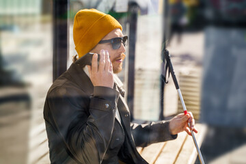 Blind man with a walking stick sits on a bench at a public bus stop uses a smartphone