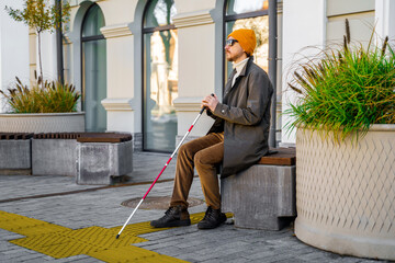 Blind man with a cane sits on a bench in the city near the tactile tiles