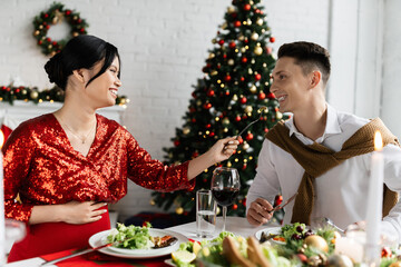 cheerful and pregnant asian woman feeding husband while having fun during romantic Christmas supper