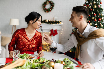 cheerful man feeding pregnant and elegant asian wife during Christmas supper at home