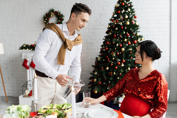 young man pouring water into glass near elegant and pregnant asian wife at romantic Christmas supper