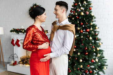 Obraz premium side view of pregnant and elegant asian woman with husband smiling at each other near Christmas tree in living room