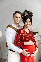 Obraz premium pregnant and elegant asian woman smiling at camera near husband hugging her belly