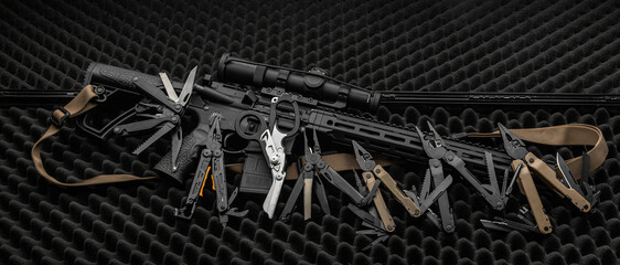 Various multi-tools lie near the automatic carbine on a gray foam surface. Dark back.
