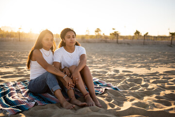 Fototapeta premium Hispanic lesbian couple sit on a blanket at the beach