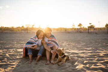 Fototapeta premium Hispanic lesbian couple sit on the beach with a blanket and dog