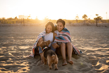 Hispanic lesbian couple sit on the beach with a blanket and dog