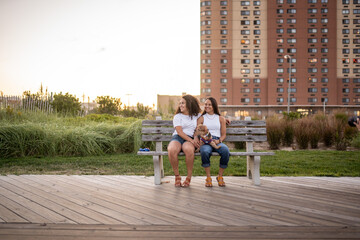 Hispanic lesbian couple sit on a bench with their dog on the boardwalk - obrazy, fototapety, plakaty