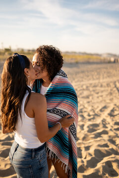 Hispanic lesbian couple kiss while wrapped in blanket at beach