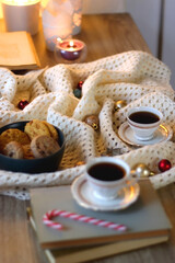 Cups of tea or coffee, bowl of cookies, soft blanket, lit candles, books and various Christmas decorations on the table. Selective focus.