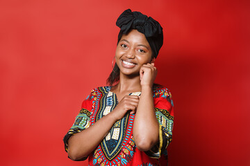 Smiling young African American woman in typical dress standing on a red background. She has her arms close to her chest as she looks at the camera