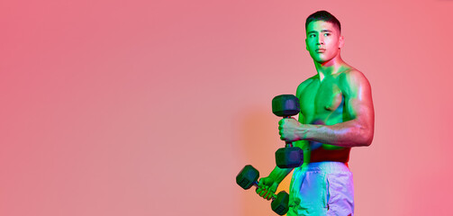 Portrait of young muscular man training, posing with dumbbells isolated over pink background in multicolored neon light.
