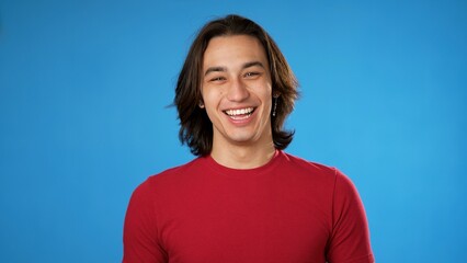Portrait of smiling laughing, happy, handsome Hispanic gender fluid young man 20s wearing red casual shirt isolated on blue color background in studio. Sincere emotions lifestyle concept.