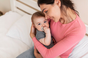 Loving mother cuddling and resting with her infant daughter on bed, mom holding cute newborn baby on hands