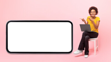 Happy young african american curly lady sit on chair with laptop, show fingers at huge smartphone with blank screen