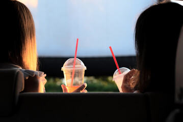 Two female friends with cocktails sitting in the car while watching a movie at drive in cinema....