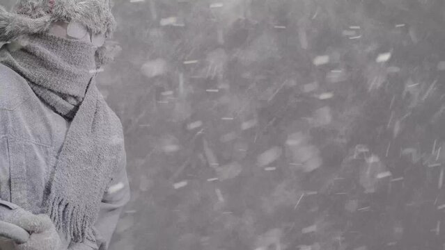 Slowly zooming out on a frozen man heading out to shovel snow all bundled up in a fur trapper&rsquo;s hat, scarf, parka, eyeglasses, covered in snow and holding a snow shovel in a blizzard.