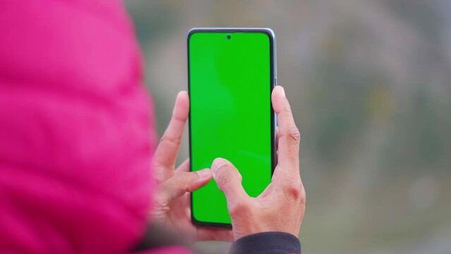 Closeup shot of Indian woman hand using mobile phone with Green screen. Green screen mockup template of Mobile phone, Chroma key. Girl wearing jacket in winter and typing in mobile phone