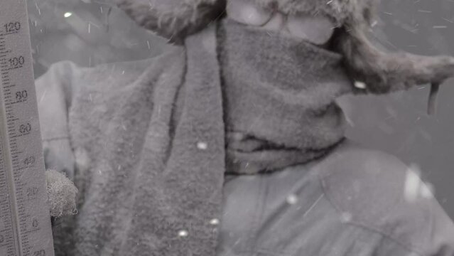 Slowly zooming out on a frozen man all bundled up in a fur trapper&rsquo;s hat, scarf, parka, eyeglasses, covered in snow and holding a thermometer in a blizzard.