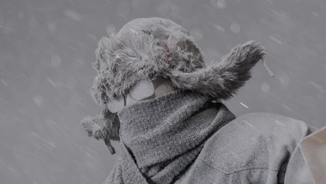 Slowly zooming out on a frozen man heading out to shovel snow all bundled up in a fur trapper&rsquo;s hat, scarf, parka, eyeglasses, covered in snow and holding a snow shovel in a blizzard.