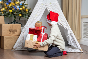 A pretty preschooler boy in a Santa hat hides his gifts in a children's tent wigwam in the nursery....