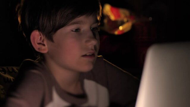 A child sits in the dark at a computer. A student studies in the dark. A little boy is watching a video on a tablet. A child emotionally watches a video. Selective soft focus