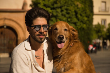 Portrait of young Hispanic man with beard, sunglasses and white shirt, sitting on a bench next to his precious dog. Concept animals, dogs, love, pets, golden.