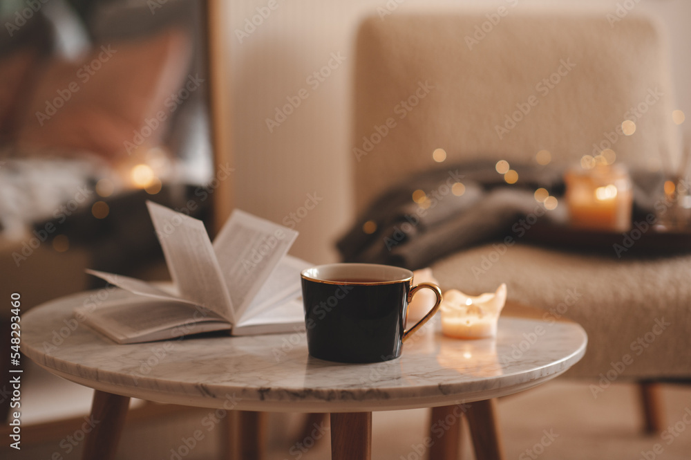 Wall mural Cup of tea with paper open book and burning scented candles on marble table over cozy chair and glowing lights in bedroom closeup. Winter holiday season.