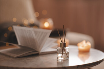 Home liquid fragrance in glass bottle and bamboo sticks with paper open book on marble coffee table over glow Christmas lights in bedroom. Winter holiday season.