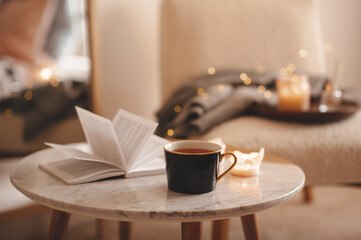 Cup of tea with paper open book and burning scented candles on marble table over cozy chair and glowing lights in bedroom closeup. Winter holiday season. - obrazy, fototapety, plakaty