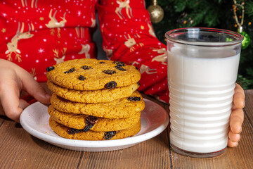 Adorable girl child in pajamas preparing cookies and milk for Santa on wooden table. Close-up on decorated Christmas tree background. Waiting for Christmas and gifts concept