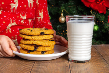 Adorable girl child in pajamas preparing cookies and milk for Santa on wooden table. Close-up on decorated Christmas tree background. Waiting for Christmas and gifts concept