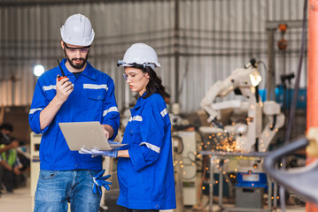 Robotic engineer male and female using laptop for workinging and controlling welding robot arm in steel factory. Maintenance system team working together with teamwork.