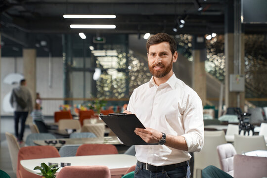 Successful entrepreneur in wearing shirt making notes, while working in modern expo center. Portrait of handsome restaurateur smiling at camera, while choosing furniture in store. Concept of business.