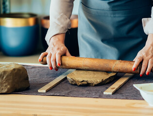 Small Business: An anonymous woman potter using rolling pin for clay mass and making ceramics object on a desk in her studio