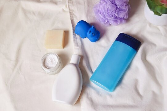 White shampoo bottle, blue shower gel package, facial cream jar, natural soap, purple sponge and rubber toy hippo. Hair and skin care cosmetics flat lay photo