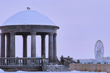 Snow-covered gazebo on the city embankment. White dome, gray granite columns and balustrade. Winter...