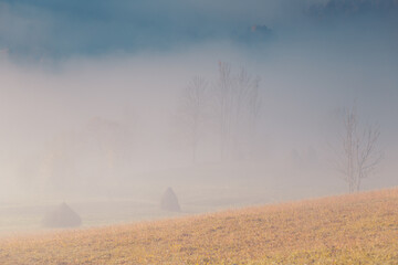 A dreamy view of the morning Carpathian mountains with sunlight through the fog in the valley. Rural scene with haystacks on pasture.