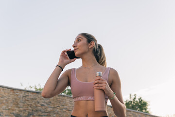 Athletic young woman holding her water bottle while making a call on mobile phone before doing sports outdoors.