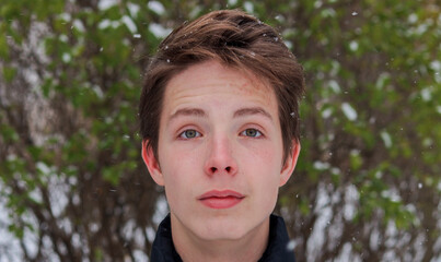 Portrait of a teenage boy close-up against the background of snow and a green bush.