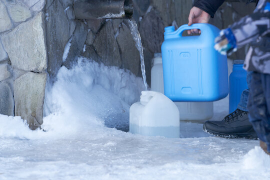 A man fills numerous containers with drinking water from a spring. Icy water pours out of a metal pipe. Plastic cans are on ice. Selective focus.