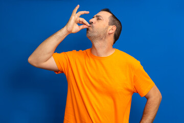 Portrait of a bearded hispanic man in orange t-shirt with puckered lips showing gourmet sign with fingers tasty delicious isolated on vibrant blue color background.