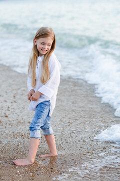 Smiling funny kid girl 5-6 year old wear white top shirt and denim pants walk in sea shore beach outdoor. Childhood. Summer season.