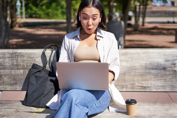 Happy asian girl chatting, having video call on laptop, sitting on bench in park and communicating, talking to someone online